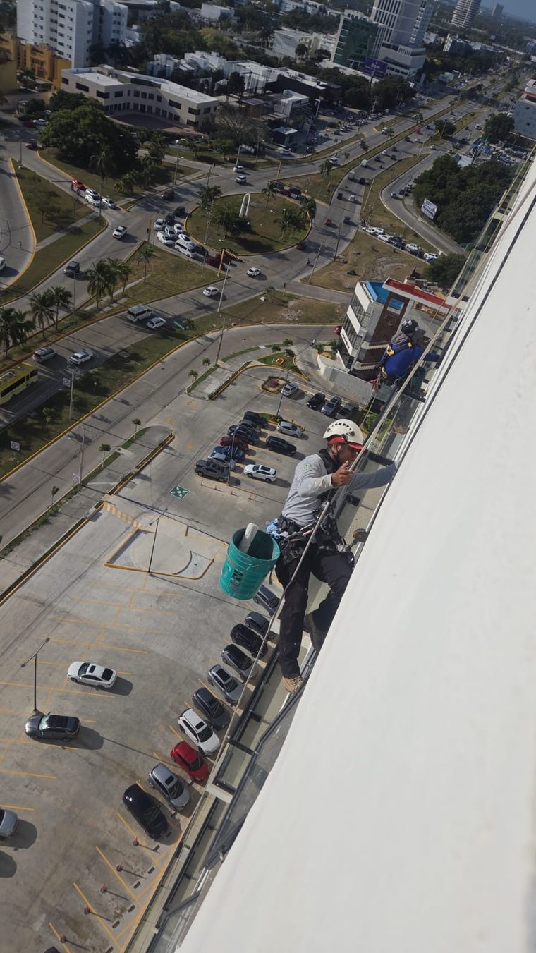 Técnico limpiando la fachada de cristal de un edificio, suspendido con arnés y casco